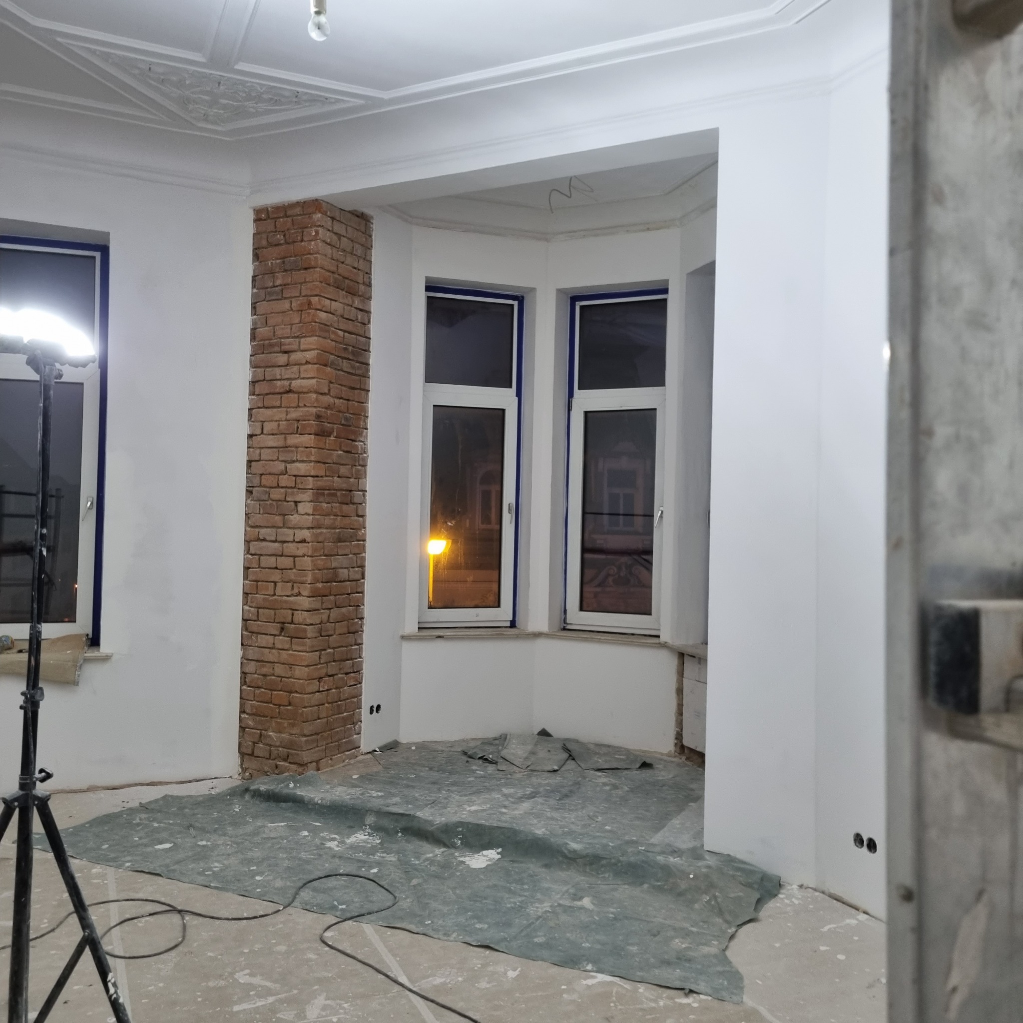 Pokój w trakcie prac wykończeniowych: surowe ściany, zabezpieczona podłoga, kolumna z cegły, widoczne okna z uliczną latarnią w tle oraz dekoracyjny sufit z ornamentami.