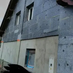 Dom w trakcie ocieplania: szare płyty styropianowe z widocznymi kołkami mocującymi pokrywają górną część elewacji, dolna część otynkowana, okno zabezpieczone folią, skrzynka elektryczna na ścianie.