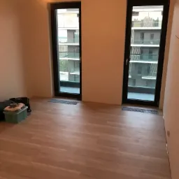 Jasna podłoga z paneli winylowych ułożona w pustym pokoju z dwoma oknami balkonowymi z czarnymi ramami, widok na budynek mieszkalny, lekki bałagan w rogu.