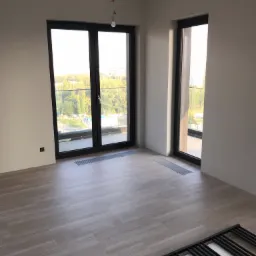 Pokój z nową podłogą winylową imitującą drewno, widok na zewnątrz przez duże okna balkonowe, grzejnik leżący na podłodze, surowa żarówka zwisająca z sufitu.