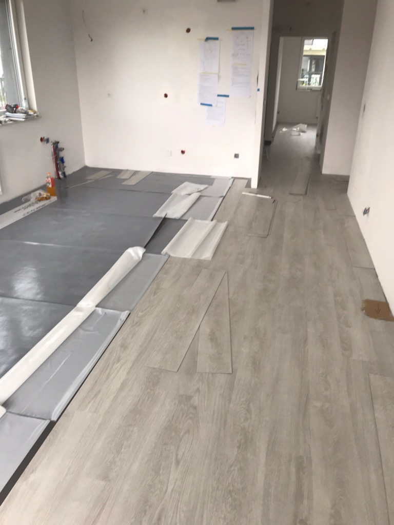 Wnętrze w trakcie montażu podłogi winylowej; widoczne rozłożone panele, podkład oraz materiały przygotowane do dalszych prac wykończeniowych.