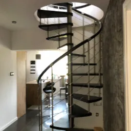 Kompaktowe schody spiralne z czarnymi stopniami i metalową balustradą w nowoczesnym wnętrzu z aneksem kuchennym w tle.