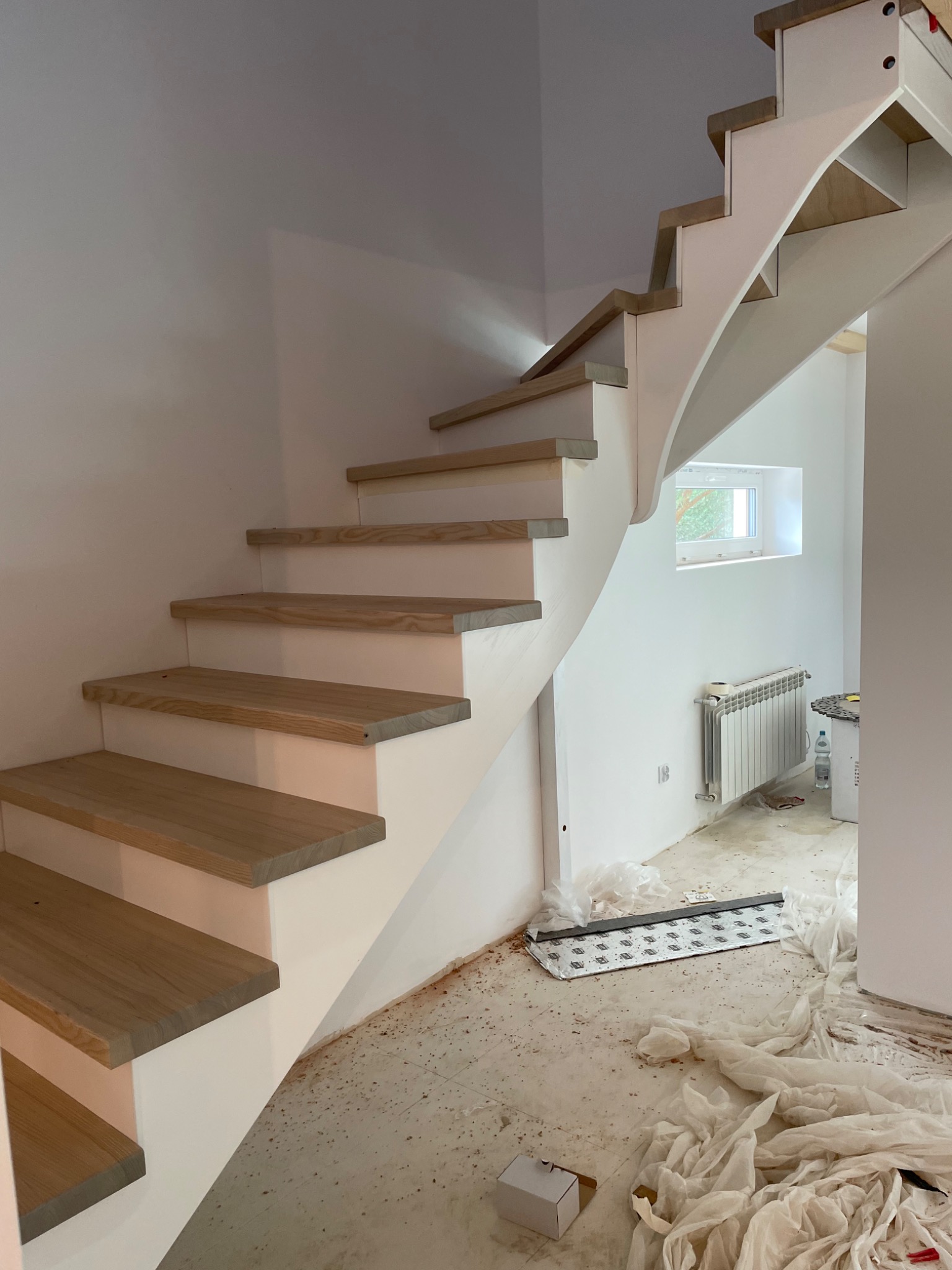 Montaż ażurowych schodów z jasnego drewna na białej konstrukcji w surowym wnętrzu, widoczne zabezpieczenia podłogi i grzejnik.