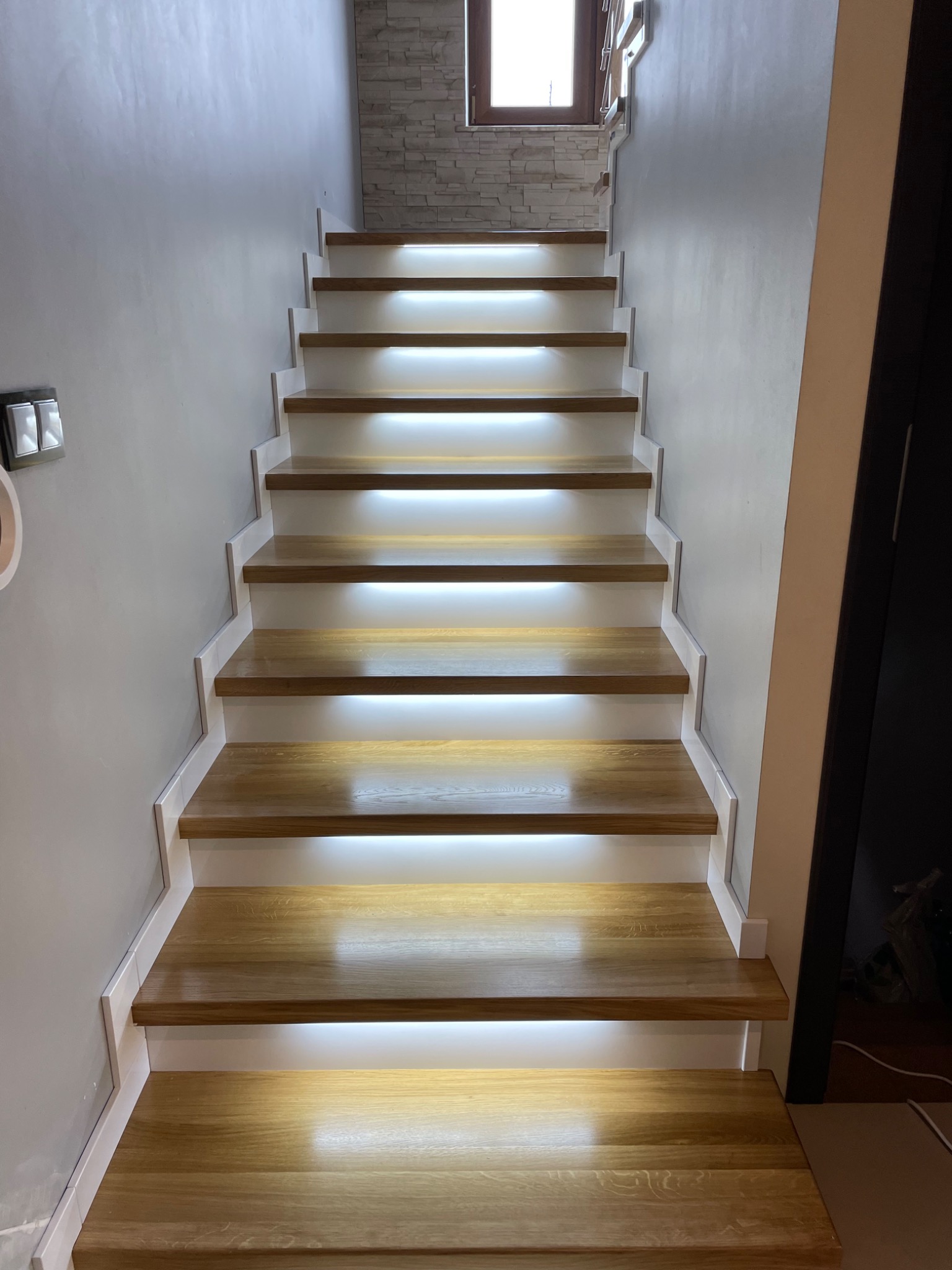Nowoczesne schody z jasnego drewna z podświetleniem LED pod każdym stopniem, widok z dołu na górę, białe ściany i fragment kamiennej ściany w tle.