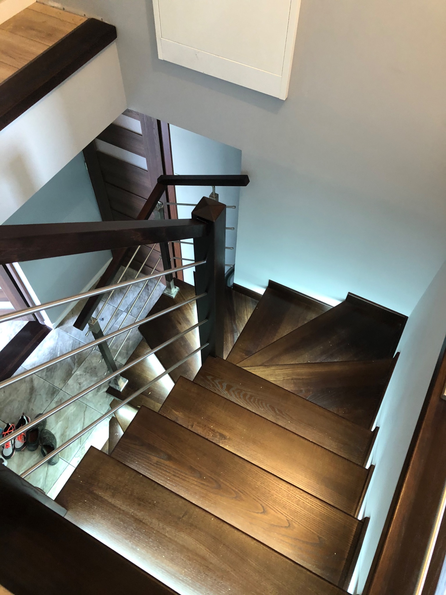 Widok z góry na ciemne, drewniane stopnie schodów z metalową balustradą w nowoczesnym wnętrzu, z widocznym fragmentem podłogi z płytek i parą butów w rogu.