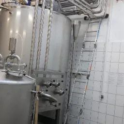 Spawacz w niebieskim kombinezonie i czapce, siedzący na szczycie dużego, metalowego zbiornika, podczas spawania rur w przemysłowym otoczeniu z białymi kafelkami i metalowymi rurami.