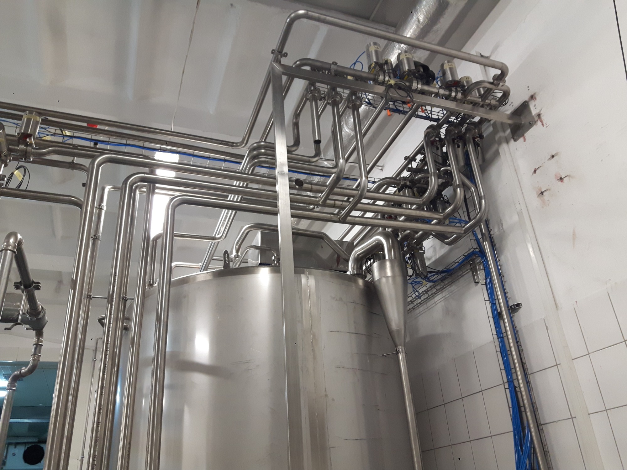 Instalacja przemysłowa z rur ze stali nierdzewnej, widoczny zbiornik i liczne rury, elementy montażowe i okablowanie, całość w pomieszczeniu z białymi płytkami.
