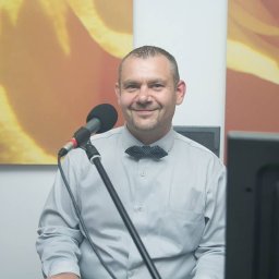 Top serwis - Uśmiechnięty mężczyzna z krótkimi włosami i muszką w kropki siedzi przed mikrofonem na tle abstrakcyjnej grafiki w odcieniach pomarańczy.