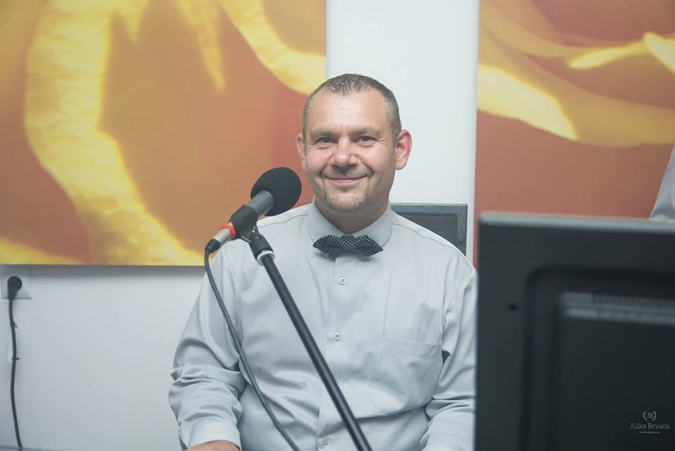 Uśmiechnięty mężczyzna z krótkimi włosami i muszką w kropki siedzi przed mikrofonem na tle abstrakcyjnej grafiki w odcieniach pomarańczy.