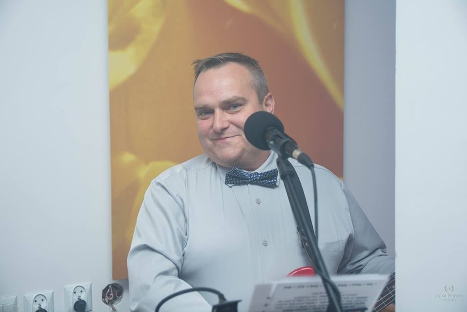 Uśmiechnięty muzyk Mariusz z niebieską muszką, śpiewa do mikrofonu, trzymając w rękach czerwony instrument przypominający ukulele, w tle abstrakcyjny pomarańczowy obraz.