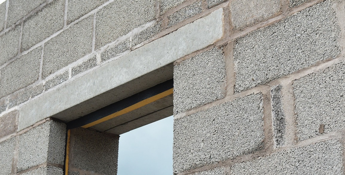 Betonowy nadproże nad otworem okiennym w ścianie z szarego betonu komórkowego, widoczne czarne elementy izolacji termicznej i żółta folia zabezpieczająca.