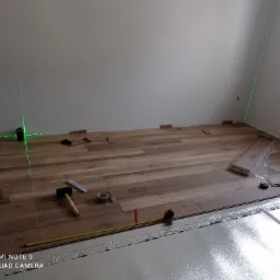 Układanie paneli podłogowych w Toruniu: widoczny laser krzyżowy wyznaczający poziom, rozłożona folia izolacyjna, częściowo ułożone panele, gumowy młotek, miarka i ołówek.