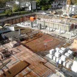 Widok z góry na plac budowy z widocznymi fundamentami, zbrojeniami, betonowymi blokami i żółtym dźwigiem w tle.