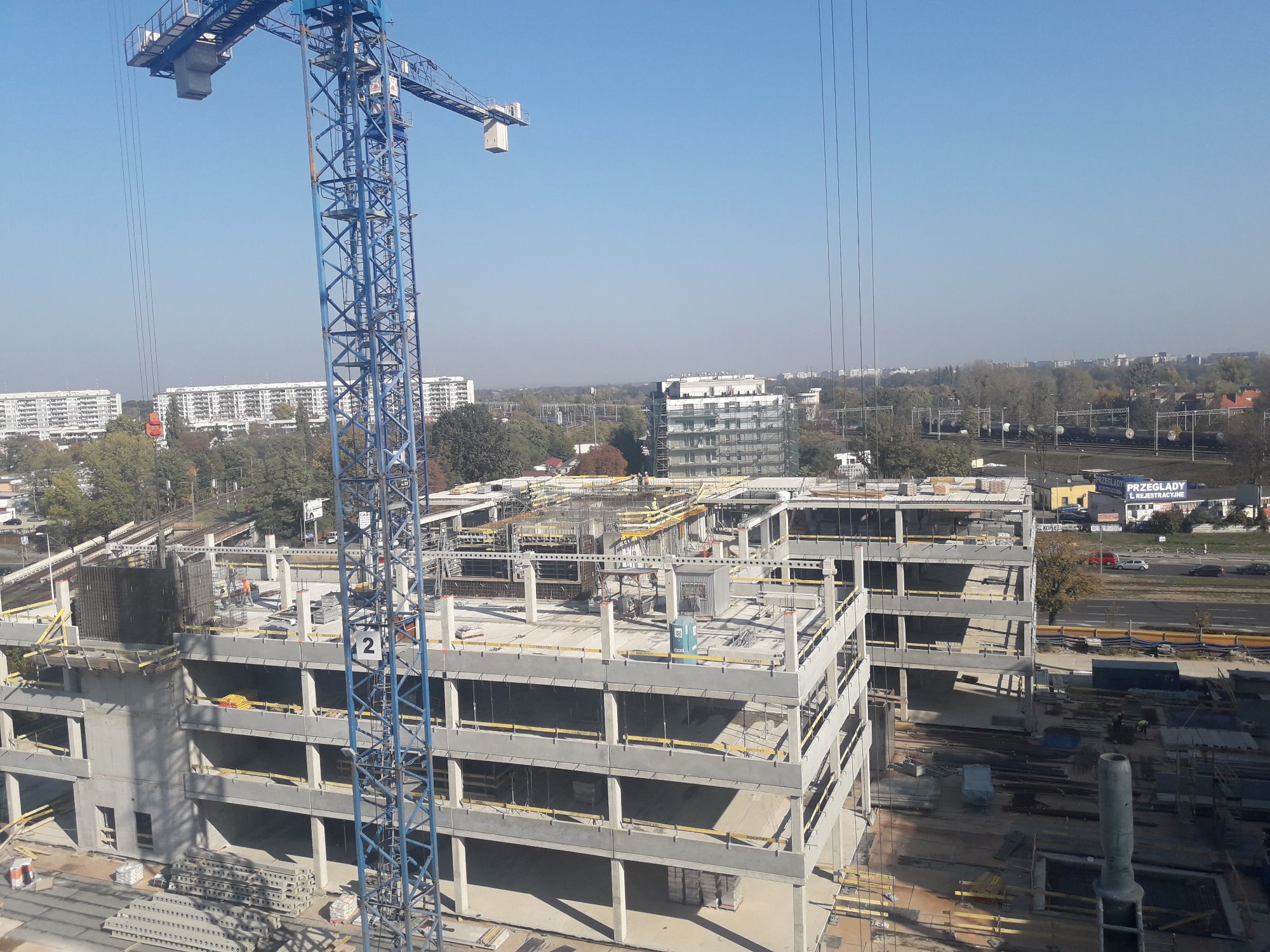 Wysoka żuraw budowlany na tle konstrukcji w stanie surowym otwartym, widok z góry na plac budowy z elementami szalunków i zbrojeń.