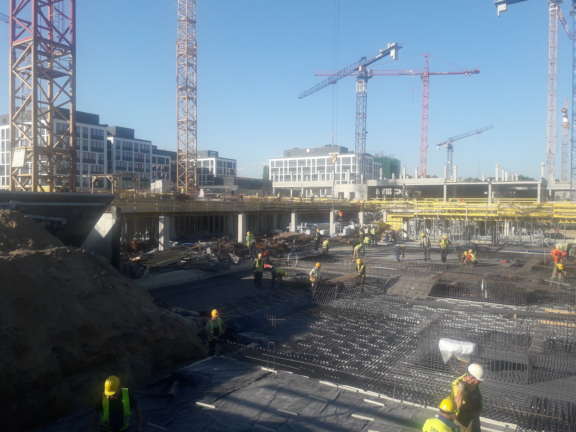 Budowa wielopiętrowego budynku w trakcie realizacji: robotnicy w żółtych kaskach układają zbrojenie na przygotowanym fundamencie; w tle widoczne żurawie i budynki.