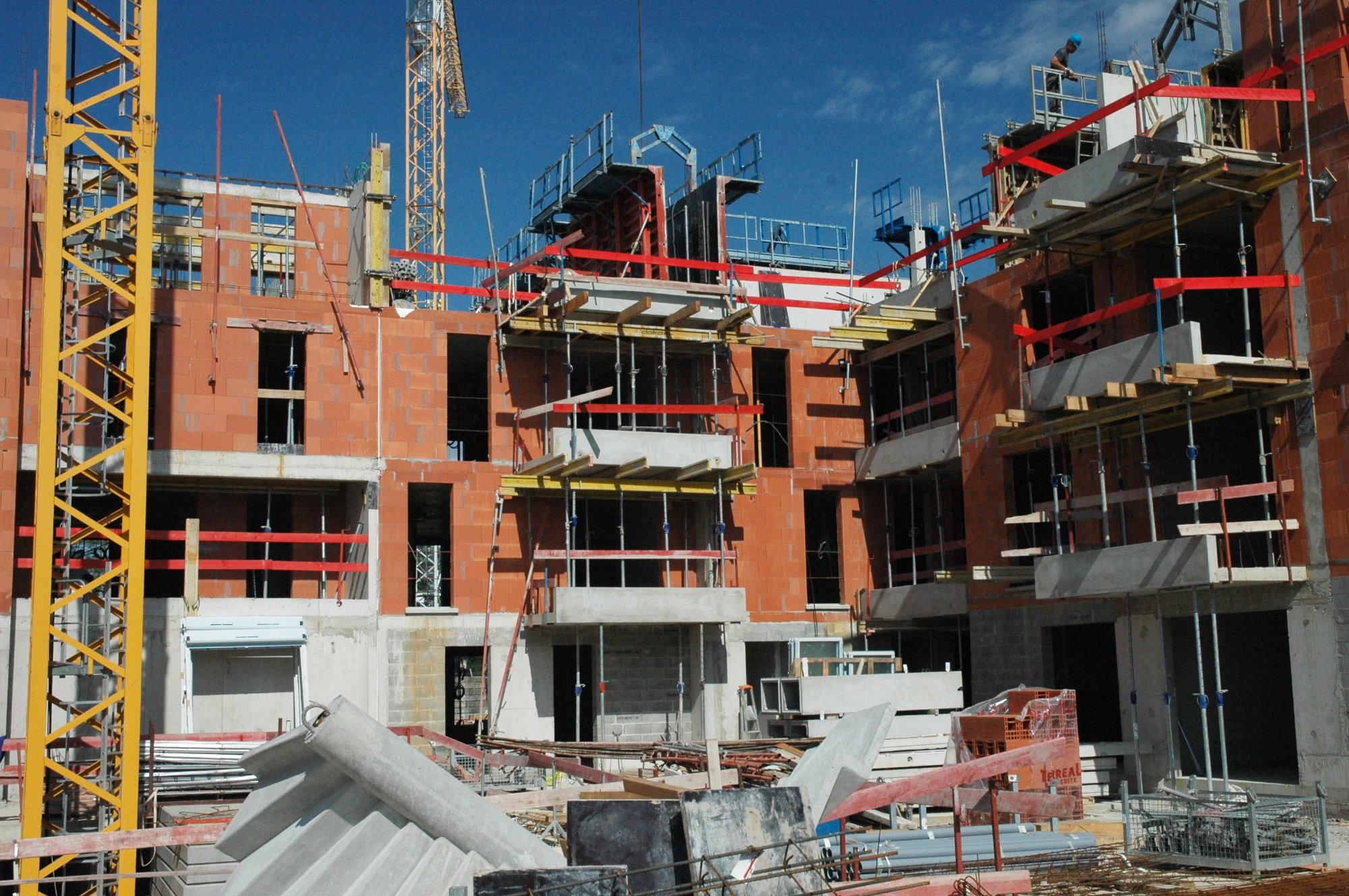 Wielopiętrowy budynek w stanie surowym otwartym, z widoczną konstrukcją z czerwonej cegły dziurawki, szalunkami, rusztowaniami i żurawiem wieżowym w tle, na pierwszym planie składowane elementy...