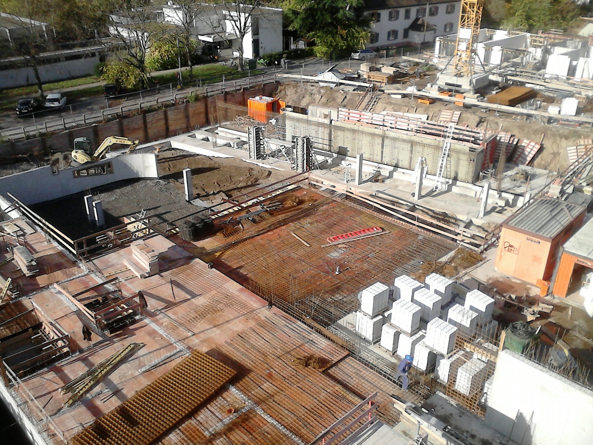 Widok z góry na plac budowy z widocznymi fundamentami, zbrojeniami, betonowymi blokami i żółtym dźwigiem w tle.