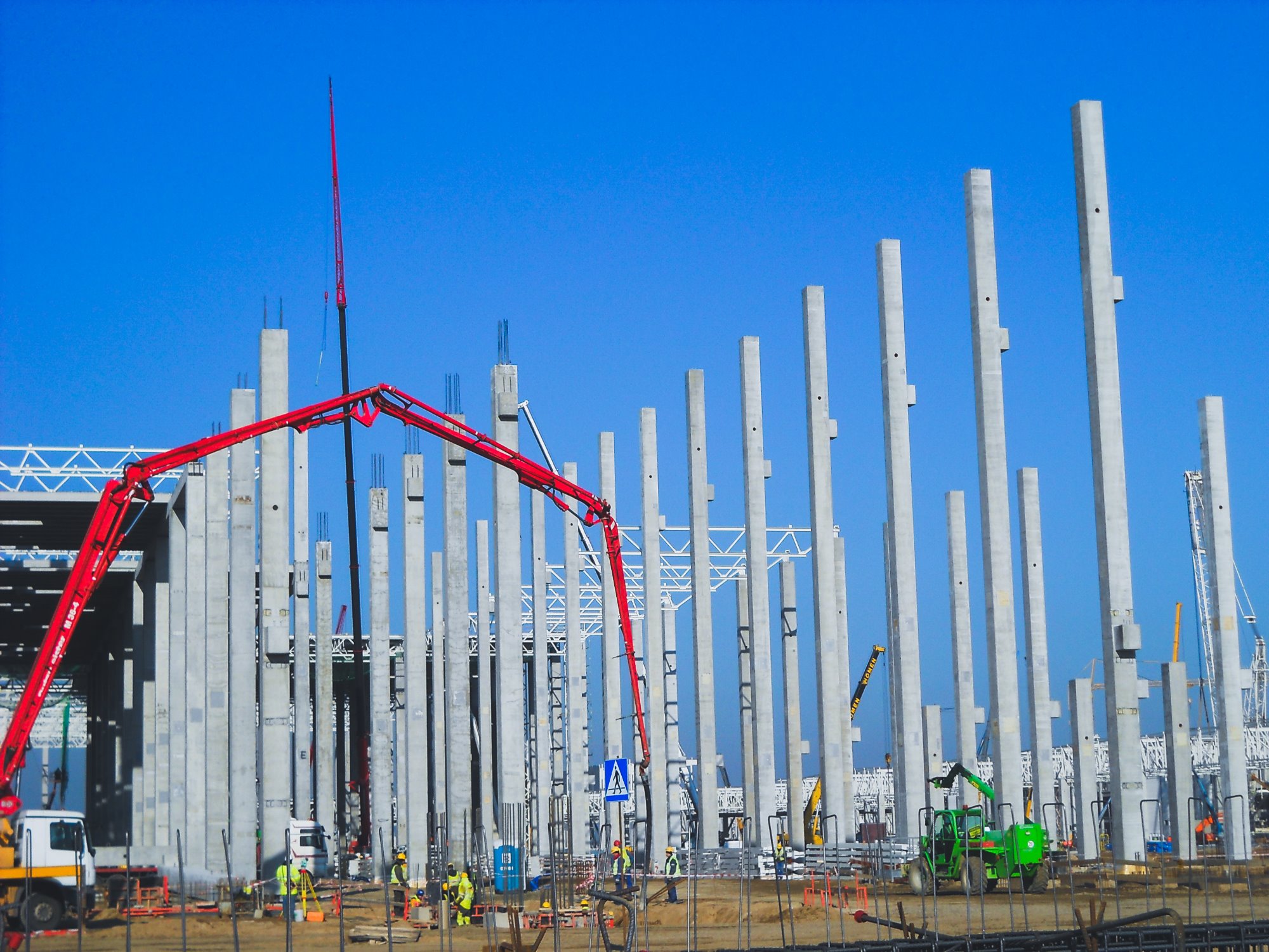 Wznoszenie konstrukcji hali: widok na betonowe słupy i dźwig do betonu na tle błękitnego nieba, pracownicy w żółtych kamizelkach w tle.