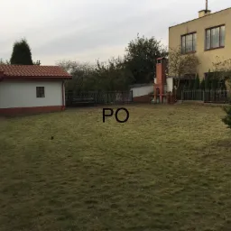 Rozległy trawnik przed domem z widocznymi budynkami w tle, świeżo skoszona trawa, częściowo zaparkowany samochód z lewej strony