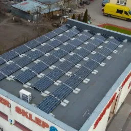 Widok z góry na dach budynku 'SUPER SKLEP' z zainstalowanymi panelami słonecznymi; na dachu widoczny także klimatyzator zewnętrzny, a w tle żółty samochód dostawczy.