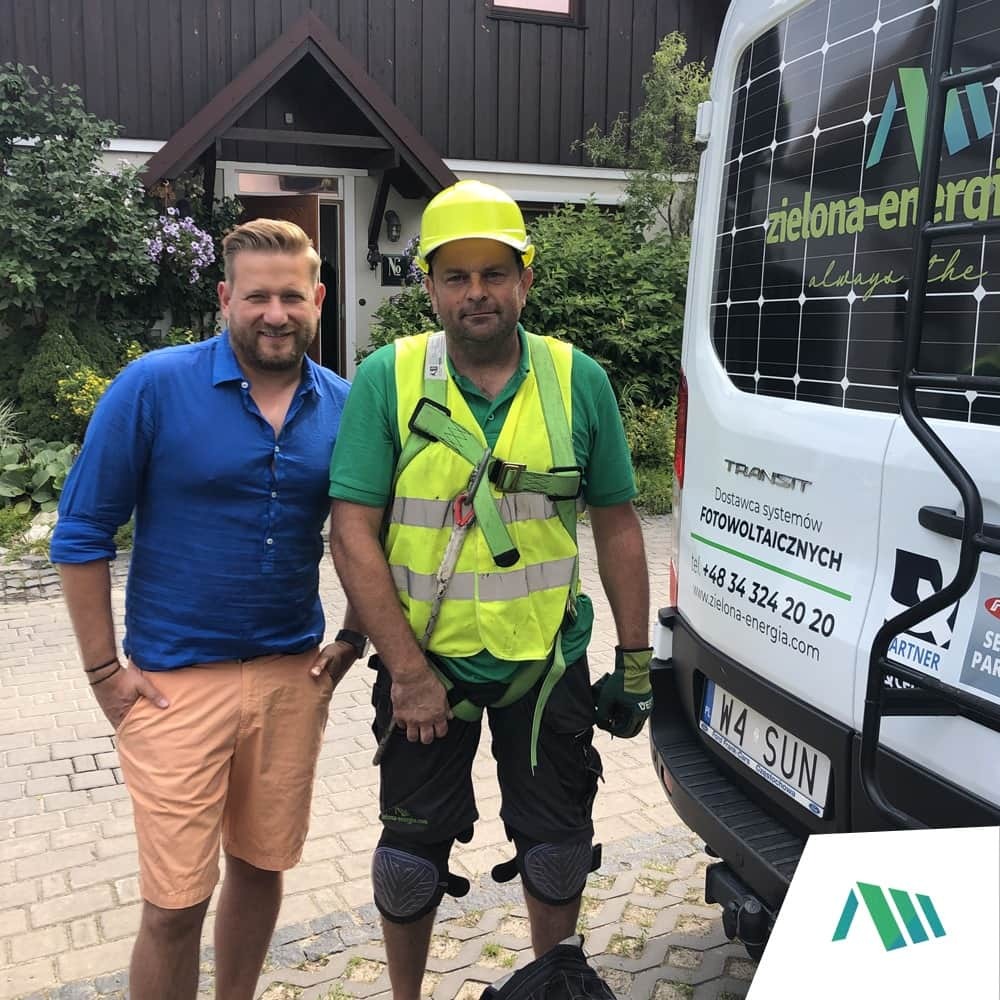 Dwóch mężczyzn pozuje obok białego furgonetki z logo firmy i informacjami kontaktowymi dotyczącymi systemów fotowoltaicznych, jeden w stroju roboczym z kaskiem i uprzężą, drugi w koszuli i szortach...