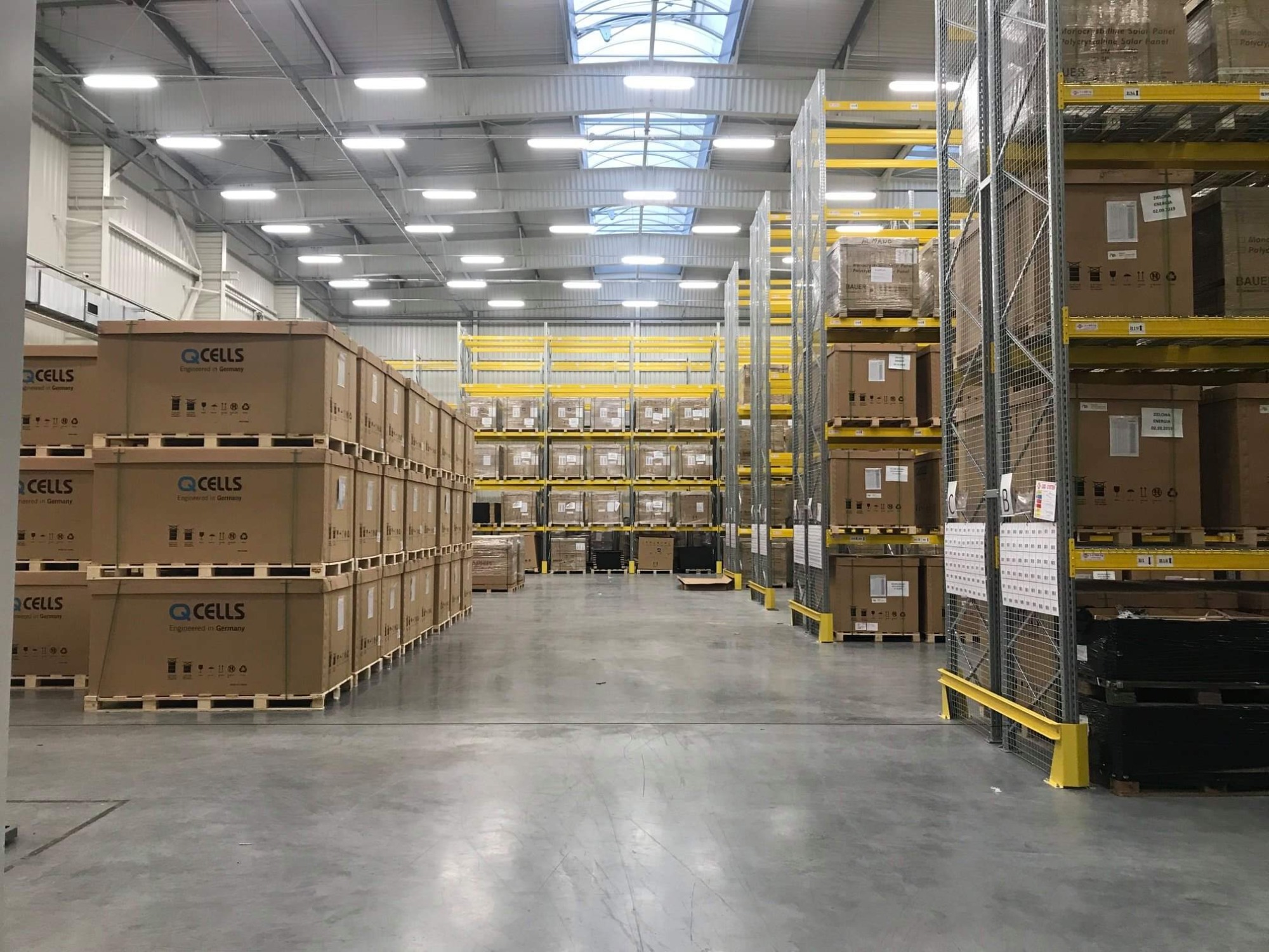Magazyn z paletami kartonowych pudeł z panelami solarnymi Q Cells ułożonymi w rzędach na żółtych regałach.
