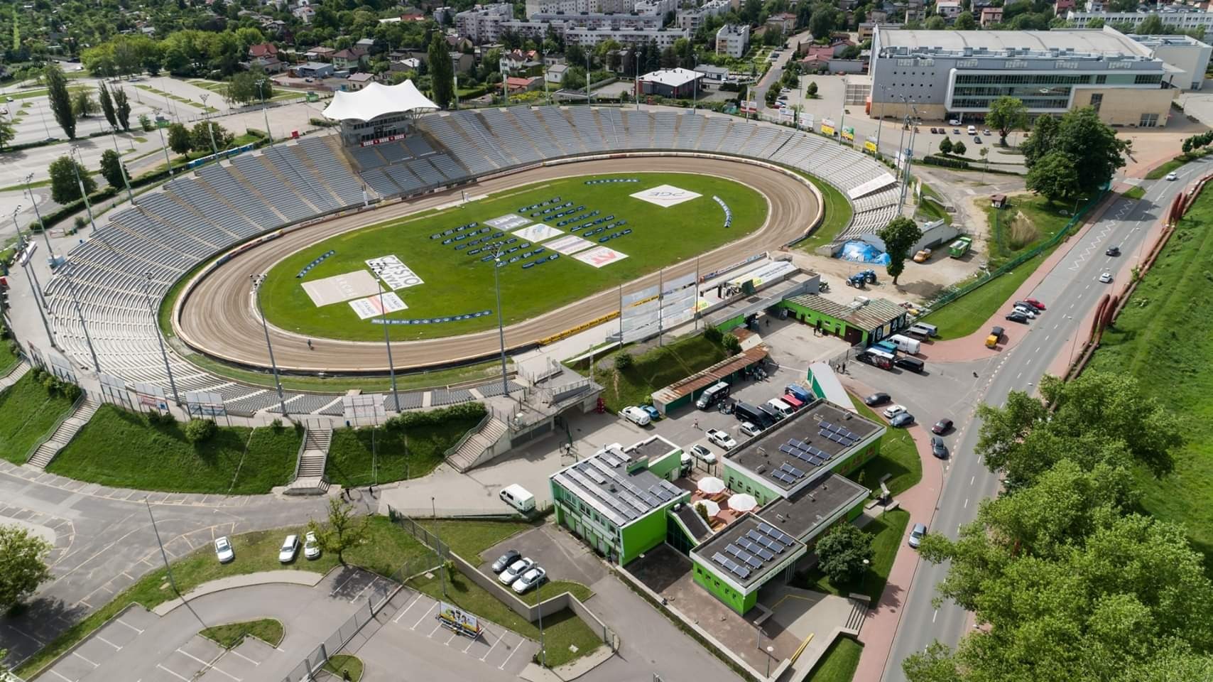 Widok z lotu ptaka na stadion żużlowy z panelami słonecznymi na dachu budynku administracyjnego, otoczony zielenią i zabudową miejską.