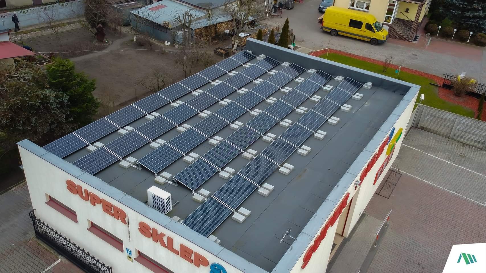 Widok z góry na dach budynku 'SUPER SKLEP' z zainstalowanymi panelami słonecznymi; na dachu widoczny także klimatyzator zewnętrzny, a w tle żółty samochód dostawczy.