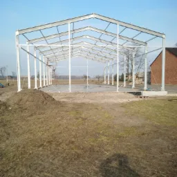 Wznoszona konstrukcja stalowa hali na betonowej wylewce, widoczny pracownik na rusztowaniu wewnątrz szkieletu, w tle ceglany budynek i błękitne niebo.