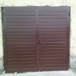 Brązowa, dwuskrzydłowa brama garażowa z poziomych paneli, zamontowana w ścianie z widocznymi uszkodzeniami i odpadającym tynkiem.