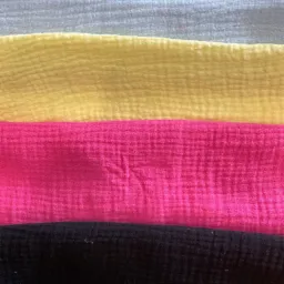 Warstwy tkaniny muslin w kolorach szarym, żółtym, różowym i czarnym, ułożone jedna na drugiej, ukazujące charakterystyczną, pofałdowaną fakturę materiału.