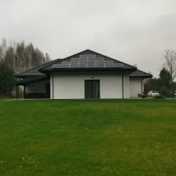 Biały dom jednorodzinny z czarnym dachem pokrytym panelami słonecznymi, widok z przodu na zadbany trawnik w pochmurny dzień.