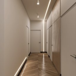 Natalia Korczyńska - Nowoczesny korytarz z parkietem w jodełkę, oświetleniem LED wzdłuż ścian i sufitu, oraz zabudową meblową z drewnianymi frontami. Minimalistyczny design.