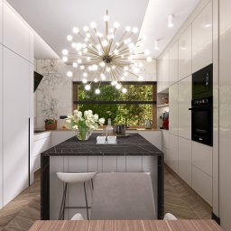 Natalia Korczyńska - Elegancka, nowoczesna kuchnia z wyspą i marmurowym blatem, oświetlona designerską lampą. Widok na okno z zielenią w tle i półki z dekoracyjnymi butelkami.