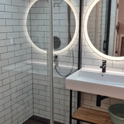Czystość - Nowoczesna łazienka z kabiną prysznicową typu walk-in, okrągłymi lustrami z podświetleniem LED, białą umywalką na czarnym stelażu i podłogą ułożoną w jodełkę.