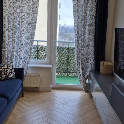 Czystość - Salon z parkietem ułożonym w jodełkę, niebieską sofą i zasłonami w kwiaty, widok na balkon z zieloną sztuczną trawą.