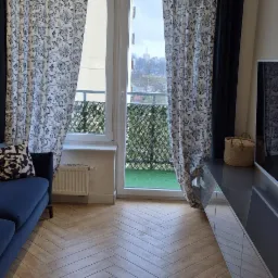 Salon z parkietem ułożonym w jodełkę, niebieską sofą i zasłonami w kwiaty, widok na balkon z zieloną sztuczną trawą.