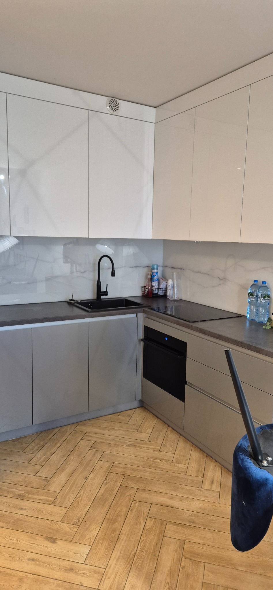 Nowoczesna kuchnia narożna z jasnymi, lakierowanymi szafkami górnymi i szarymi dolnymi, czarną armaturą i blatem, oraz podłogą ułożoną w jodełkę.