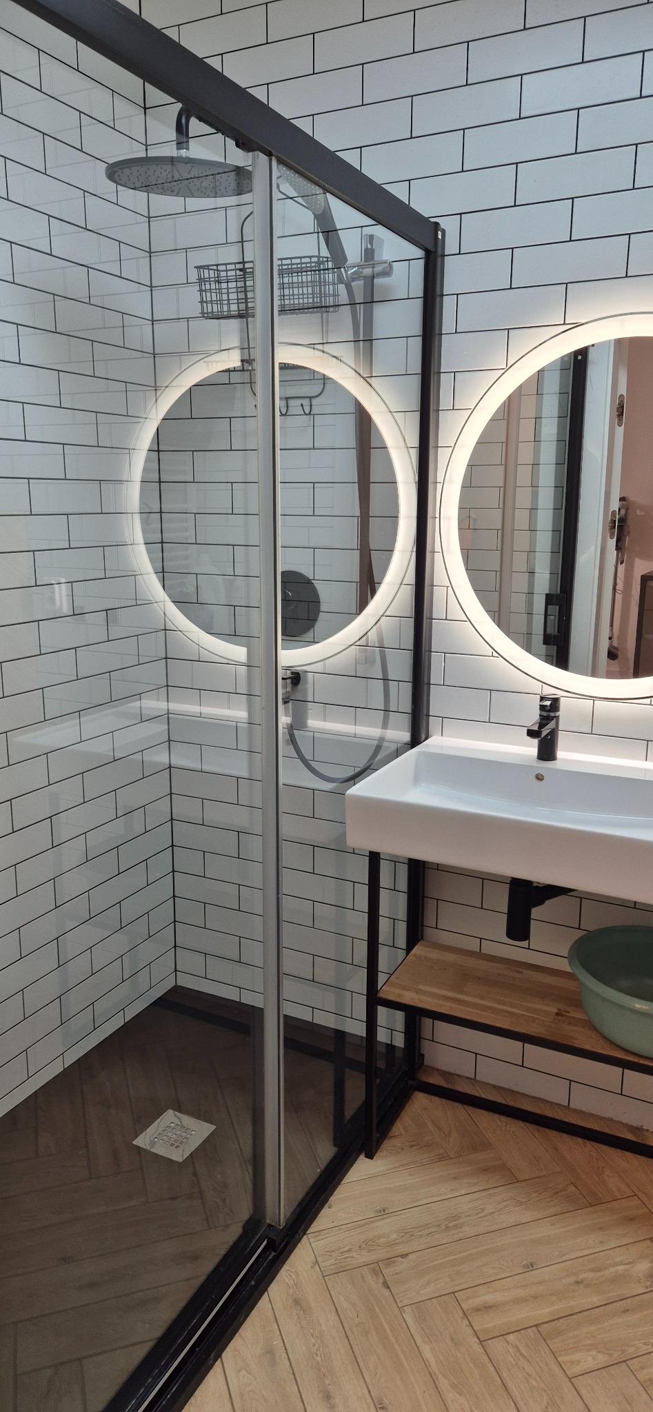 Nowoczesna łazienka z kabiną prysznicową typu walk-in, okrągłymi lustrami z podświetleniem LED, białą umywalką na czarnym stelażu i podłogą ułożoną w jodełkę.