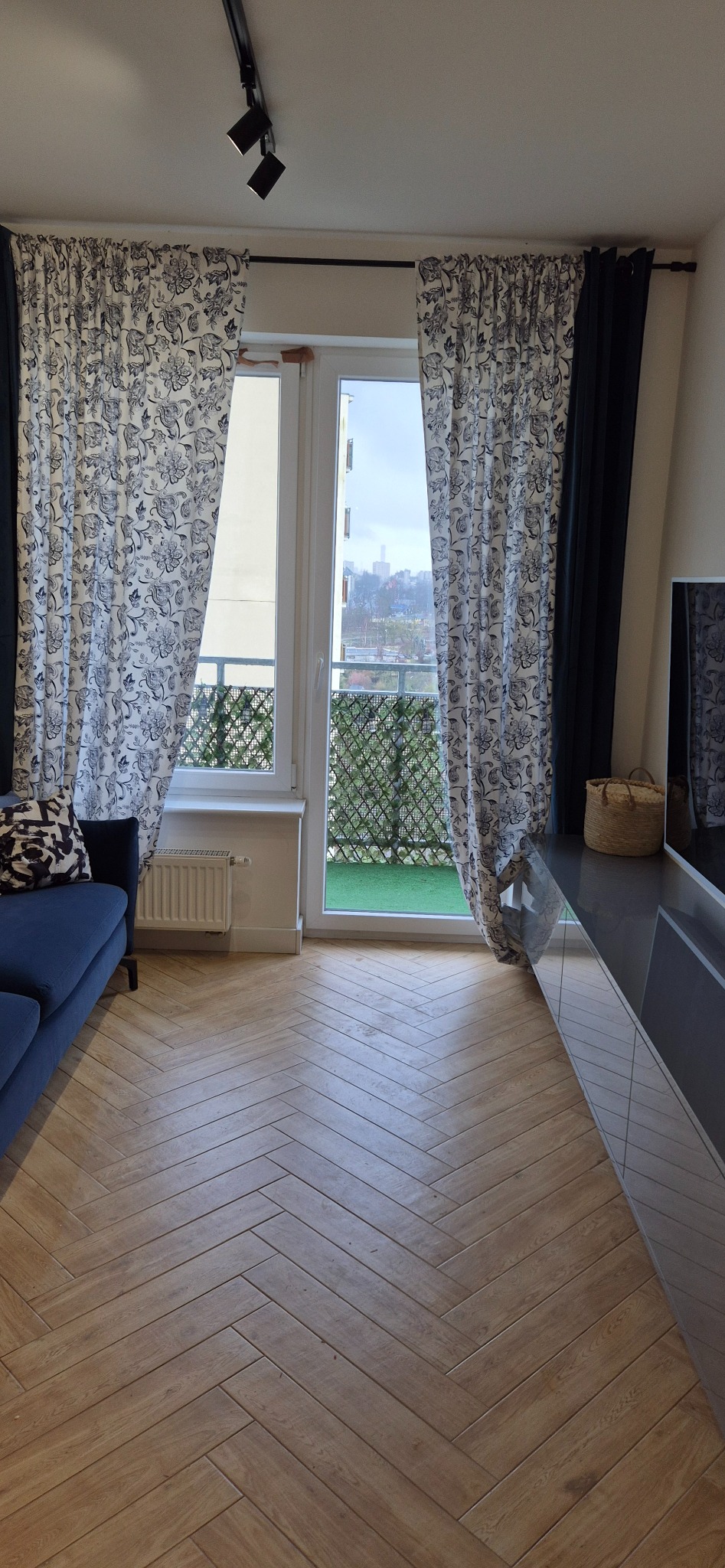 Salon z parkietem ułożonym w jodełkę, niebieską sofą i zasłonami w kwiaty, widok na balkon z zieloną sztuczną trawą.