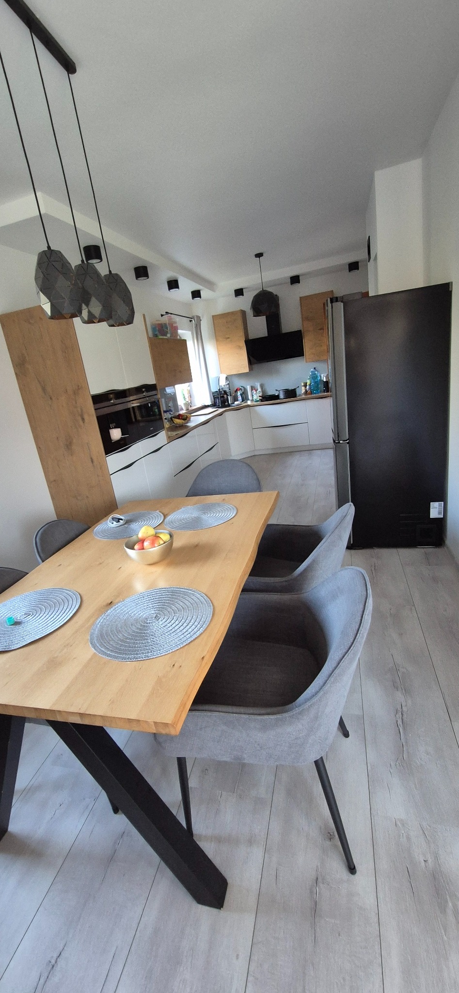 Jasne wnętrze kuchni z jadalnią, drewniany stół z szarymi podkładkami i owocami w metalowej misie, szare krzesła, nowoczesne oświetlenie i zabudowa kuchenna w tle.