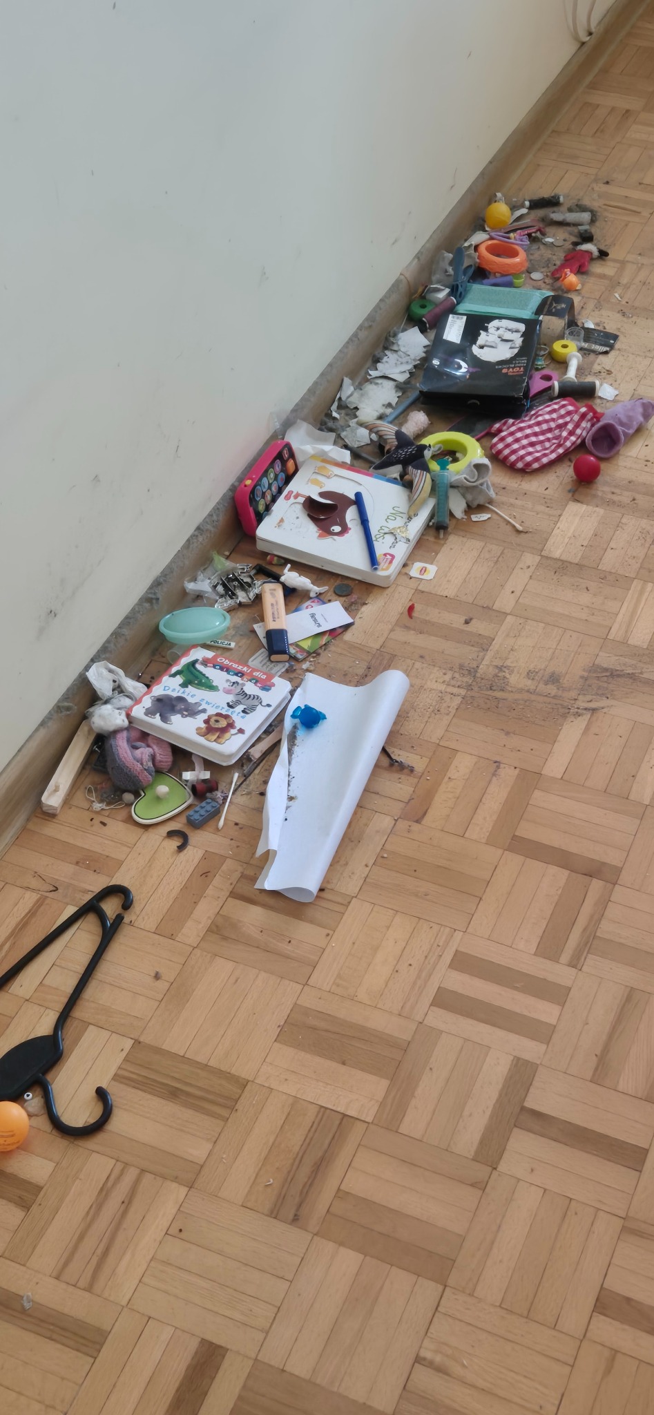 Bałagan na podłodze z parkietu: zabawki, książeczki dla dzieci, ubrania, śmieci i inne przedmioty rozrzucone wzdłuż ściany.