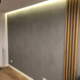 Szara ściana z dekoracyjnymi drewnianymi listwami pionowymi, oświetlona listwą LED pod sufitem, widoczny fragment podłogi z paneli i grzejnik.