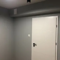 Pomieszczenie po remoncie: Ściany pomalowane na szaro, białe drzwi z czarną klamką, włącznik światła na ścianie, oświetlenie sufitowe punktowe.