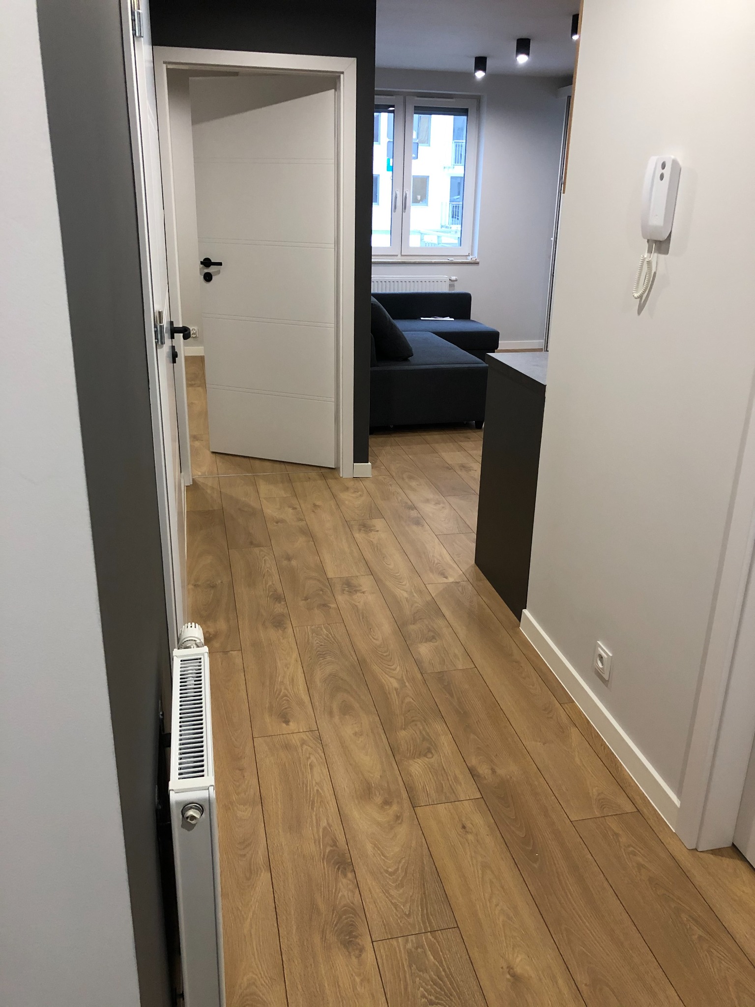 Wnętrze mieszkania po remoncie: drewnopodobna podłoga, białe drzwi z czarną klamką, szara ściana, biały grzejnik i widok na salon z szarą sofą.