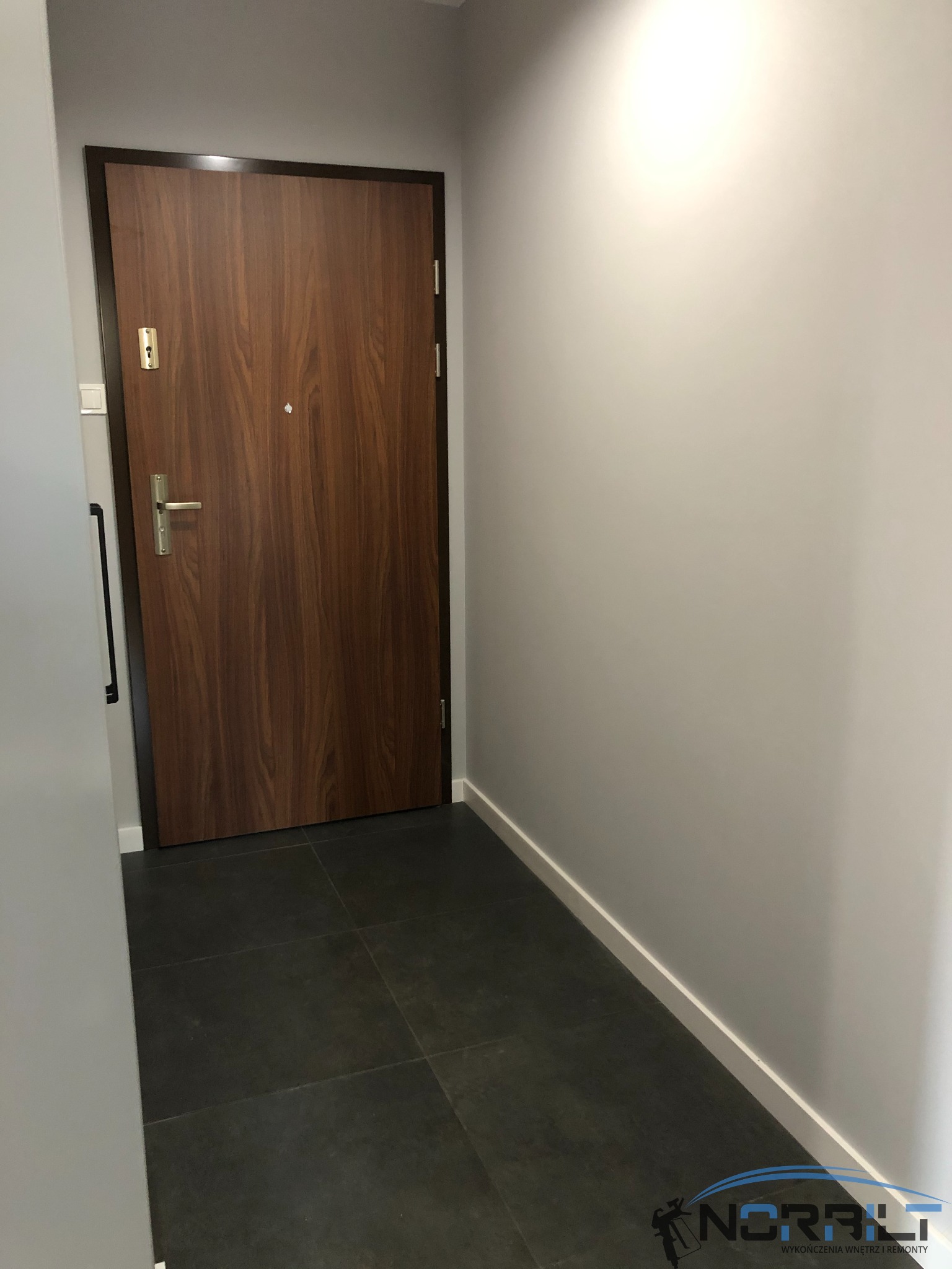 Zamknięte, drewniane drzwi wejściowe z metalową klamką i wizjerem, w szarej ścianie, ciemna podłoga z płytek, biała listwa przypodłogowa.