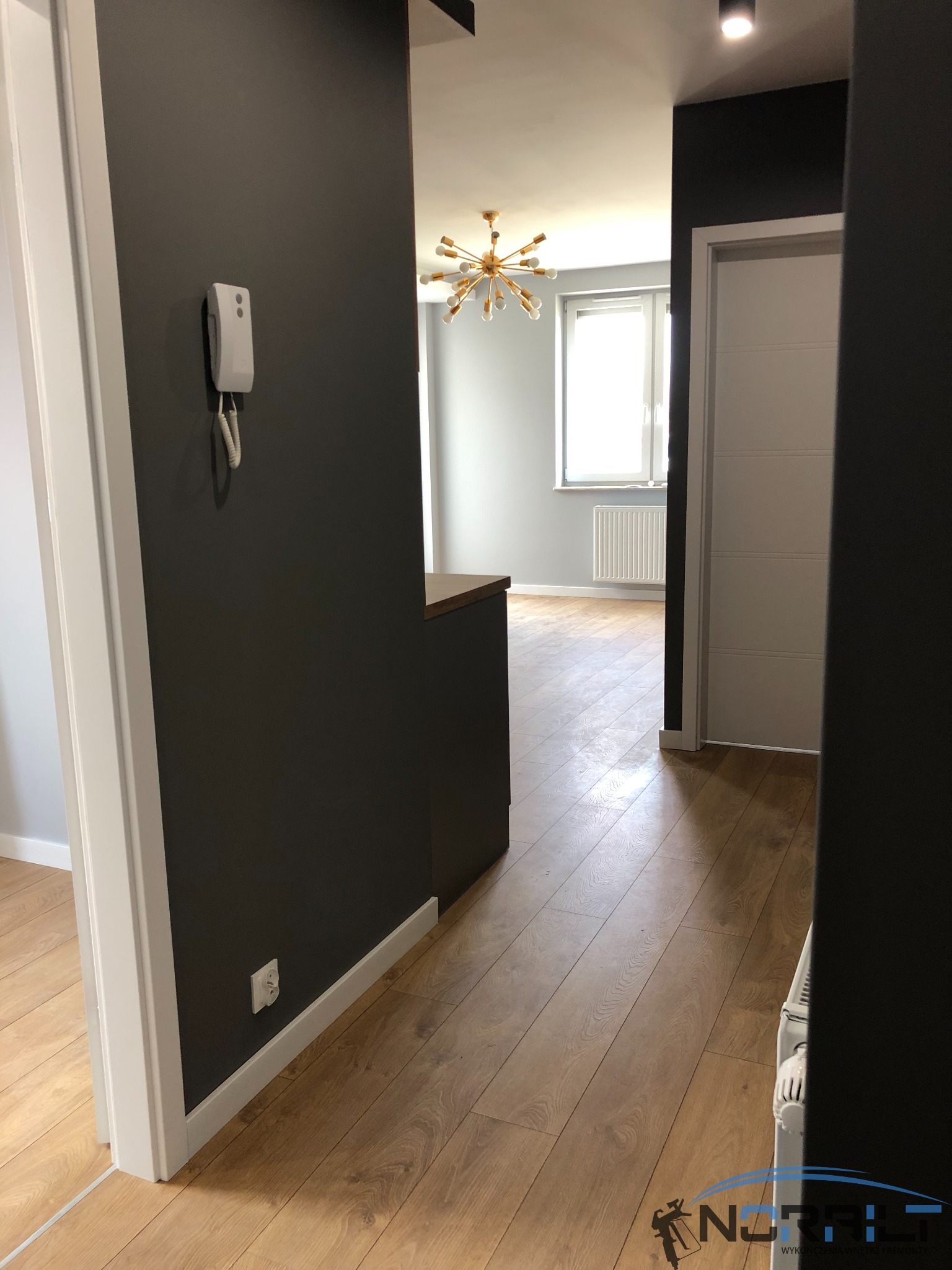 Korytarz po odświeżeniu: ciemnoszare ściany, jasne drewniane podłogi, biała stolarka drzwiowa i widoczny fragment salonu z nowoczesną lampą.