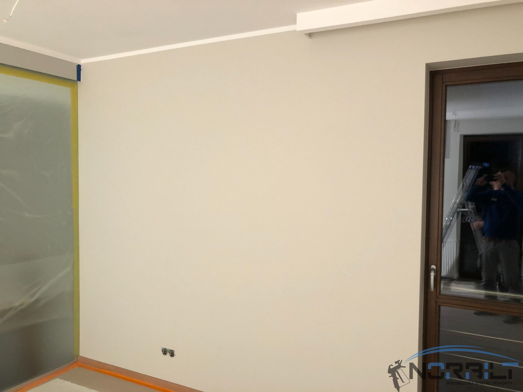 Świeżo pomalowana ściana w jasnym odcieniu z widocznymi zabezpieczeniami folią i taśmą malarską, widoczne gniazdka elektryczne, drzwi z ciemną ramą i odbicie osoby z drabiną