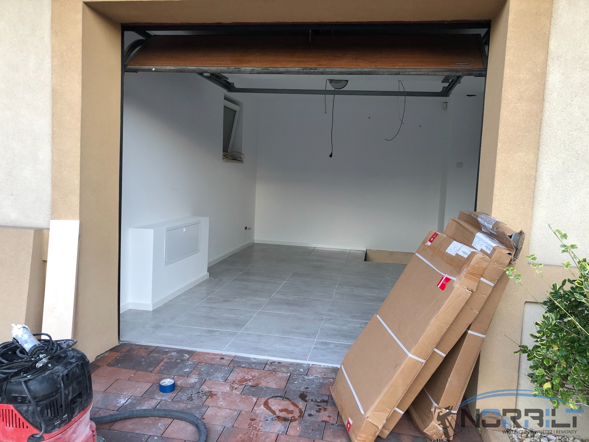 Wnętrze garażu po remoncie z szarymi płytkami podłogowymi, białymi ścianami i nową skrzynką elektryczną. Na zewnątrz leżą kartony i odkurzacz przemysłowy.