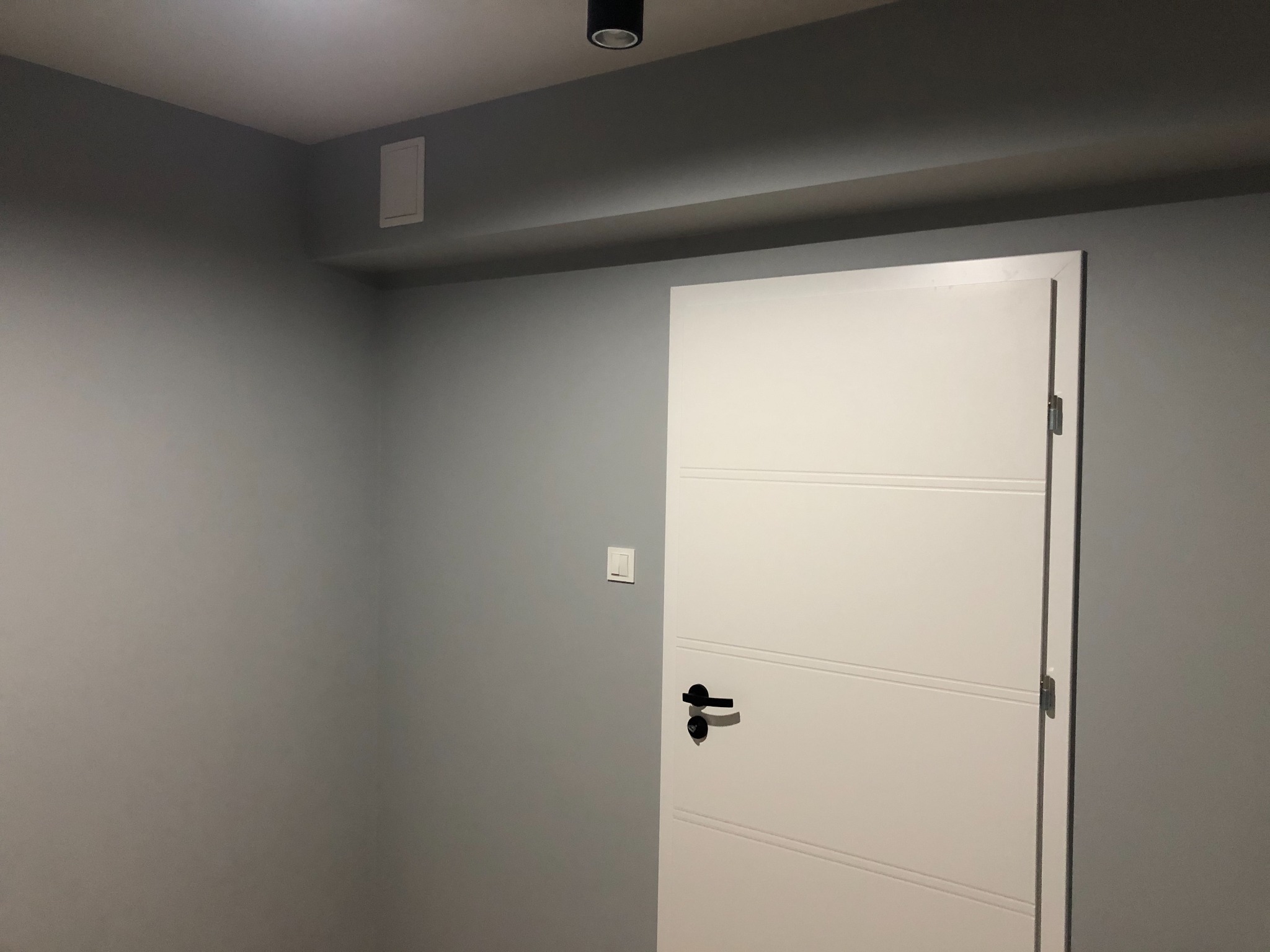 Pomieszczenie po remoncie: Ściany pomalowane na szaro, białe drzwi z czarną klamką, włącznik światła na ścianie, oświetlenie sufitowe punktowe.